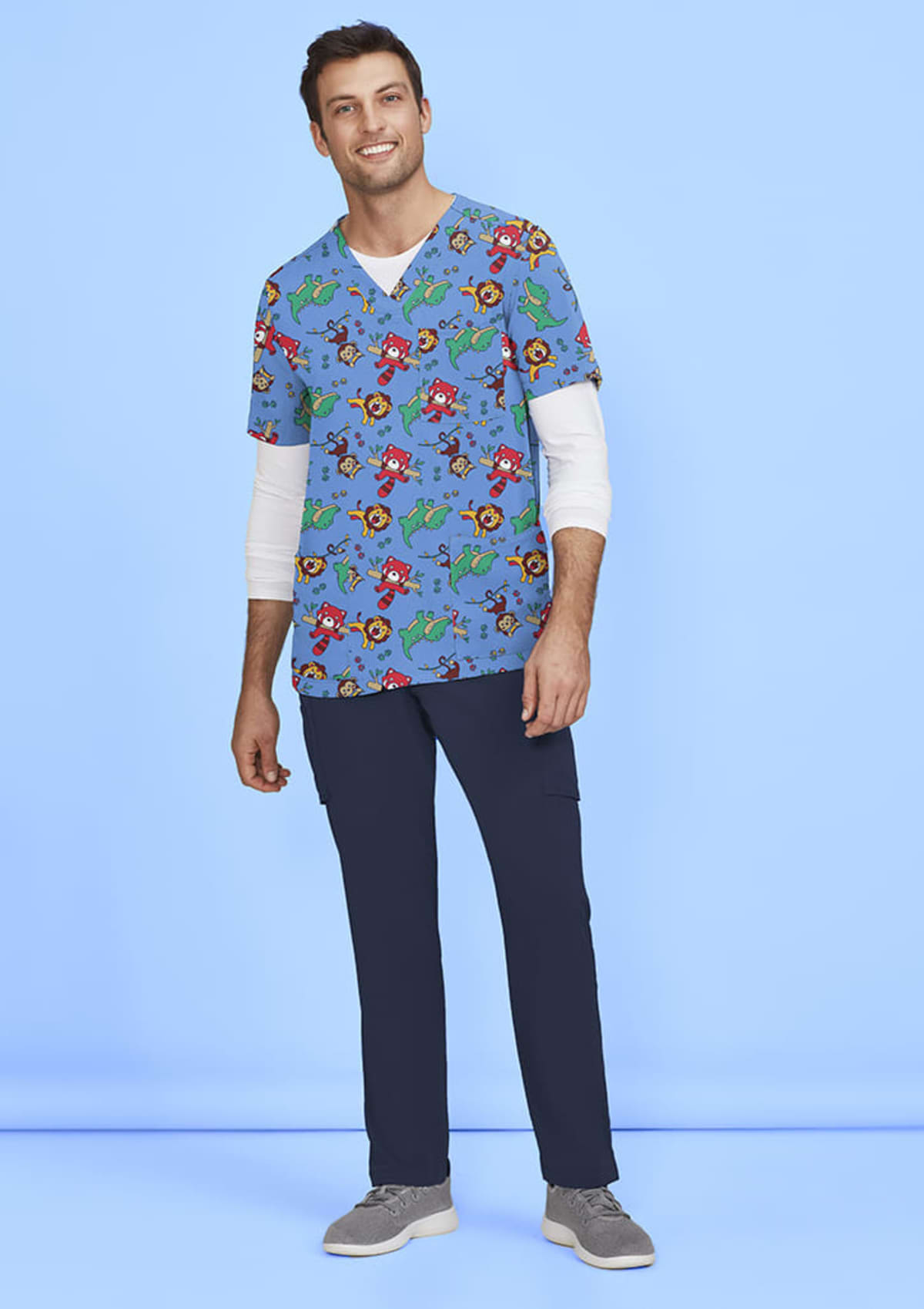 A man is standing against a light blue background, wearing colorful scrubs featuring cartoon characters and dinosaurs. He has a long-sleeve white shirt underneath and is also wearing navy blue pants and gray sneakers.