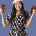 Smiling person wearing a festive reindeer antler hat and a scrub top with gnome patterns. They hold red tinsel pom-poms in both hands. The background is a solid light blue.