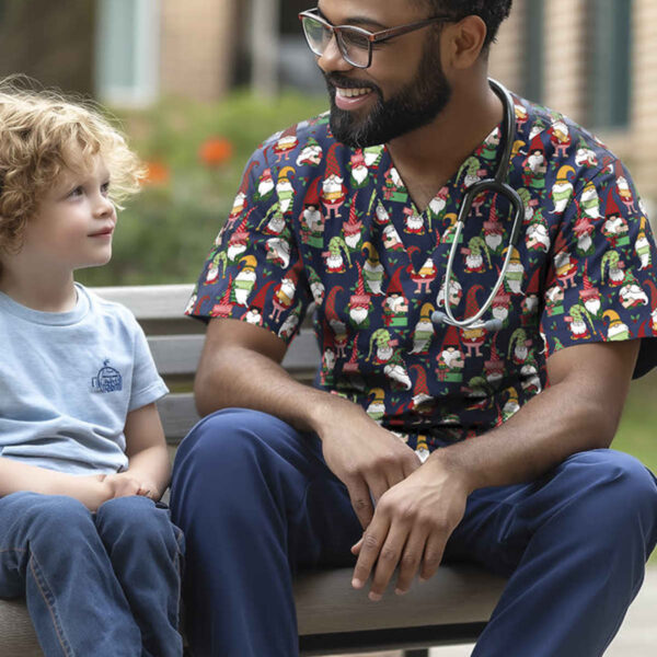 A smiling healthcare professional in festive scrubs with snowman patterns sits on a bench, engaging with a young child wearing a blue shirt. They are outdoors, with greenery and a building in the background.