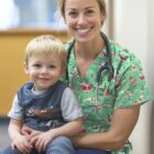 A smiling healthcare worker in green scrubs with a stethoscope around her neck holds a cheerful young boy on her lap. They are indoors, and both appear happy and relaxed.