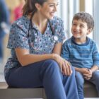 A smiling healthcare professional in scrubs with a stethoscope is sitting on a bench, talking cheerfully with a young boy in a striped shirt. They appear to be in a bright, well-lit corridor.
