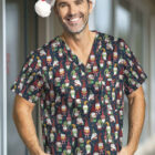 Smiling man wearing a festive Santa hat and a scrub top with colorful Santa Claus prints stands indoors with hands in pockets.
