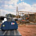 A dark blue bag with pockets containing a water bottle and various items sits on a metal surface. In the background, theres a construction site with scaffolding, wooden beams, and machinery under a partly cloudy sky.