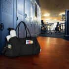 A black gym bag is placed on a wooden floor in front of a row of blue lockers. The bag contains water bottles and magazines. In the background, gym equipment is visible in a well-lit room.