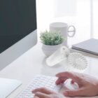 A person types on a white keyboard at a desk with a computer monitor, a small plant, a white mug, and two portable fans. The background shows a sheer curtain with soft natural light.