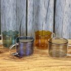 Four colored glass mugs—green, blue, amber, and clear—on a wooden surface with a wood backdrop.