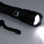 A black flashlight with a textured grip and control button emitting a bright white light. Blue indicator lights are near the button. A black lanyard is attached to its end. The flashlight is resting on a light gray surface.