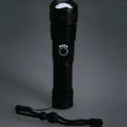 A black flashlight with a textured grip and a strap lies on a dark surface. The flashlight is standing upright with its front facing up, and its power button is illuminated, casting a small halo of light.