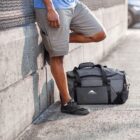 A person in a blue shirt and beige shorts leans against a textured wall with one leg bent, wearing black sneakers. Next to them is a large gray and black duffel bag on the ground.