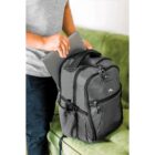 A person wearing a gray shirt is placing a laptop into a gray backpack with multiple compartments. The backpack is on a green couch.