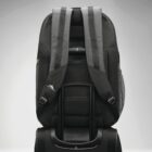 A black backpack is strapped onto the extended handle of a rolling suitcase. The backpack features padded straps and a mesh back for ventilation. The setting is a neutral, light gray background.