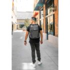 A person in a black t-shirt, dark jeans, and white sneakers walks down a sunlit urban street, carrying a gray backpack. The scene features brick buildings, storefronts, and string lights overhead.