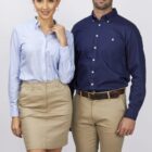 A woman and a man pose in business attire. The woman wears a light blue blouse and beige skirt. The man wears a dark blue button-up shirt and beige pants. They stand on a plain background, both smiling.