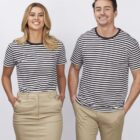 A woman and a man stand side by side, both smiling and wearing black and white striped shirts. The woman is wearing a beige skirt and the man is wearing beige pants. They both have their hands in their pockets against a plain background.