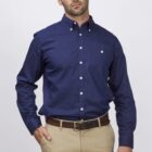 A man with short dark hair is wearing a navy blue button-up shirt and beige pants. He stands against a plain white backdrop, with one hand in his pocket and a neutral expression.