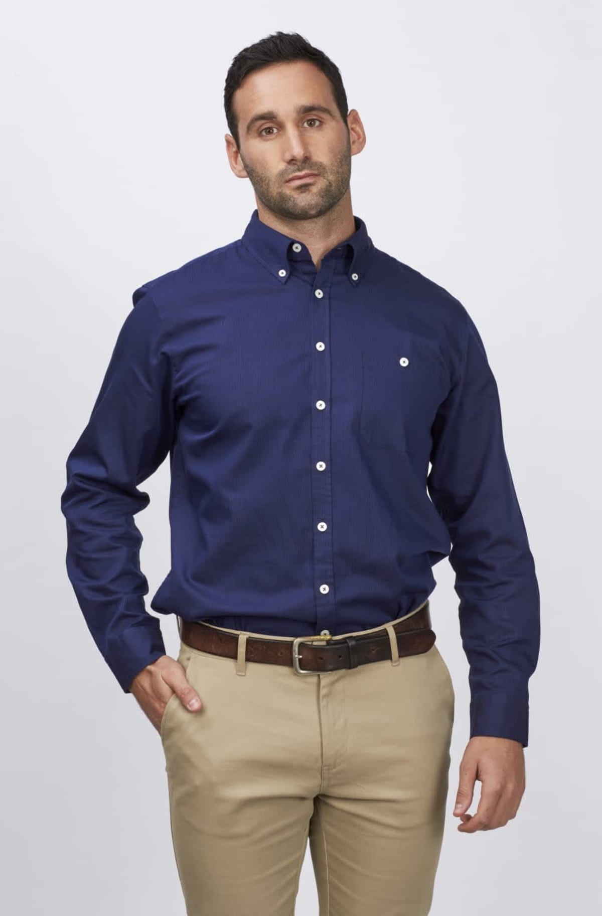A man with short dark hair is wearing a navy blue button-up shirt and beige pants. He stands against a plain white backdrop, with one hand in his pocket and a neutral expression.