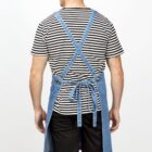 A man is standing with his back to the camera. He is wearing a striped black and white T-shirt, black shorts, and a blue apron tied at the back. The apron straps cross over his back. The background is plain white.