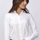 A woman in a white button-up shirt and dark pants stands holding a closed silver laptop. She has long, dark hair and is facing slightly to the side, against a plain light background.