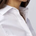 Close-up of a woman wearing a crisp white button-up shirt with a pointed collar. Her long, dark hair is visible on her shoulder, and she has a subtle, natural smile. The focus is on the shirts fabric and design.