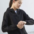 A woman with long hair tied back is outdoors, wearing a black zip-up hooded jacket. She is looking down and adjusting the sleeve on her jacket. The background is a plain light gray wall.
