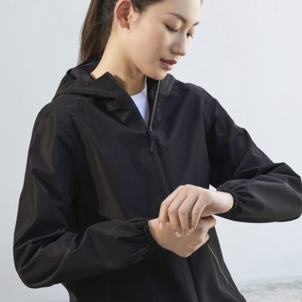 A person wearing a black hooded jacket looks down at their wrist, possibly checking a watch or wristband. The background is a simple, light-colored wall. The person has long hair tied back in a ponytail.