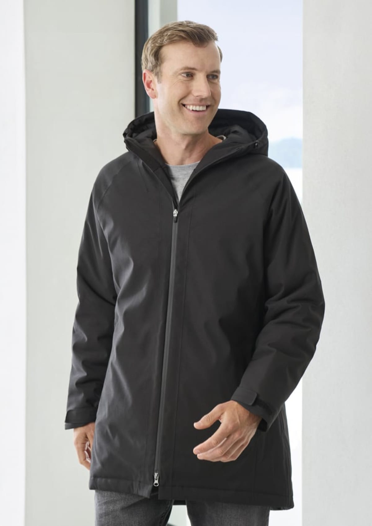 Smiling person wearing a dark hooded coat over a light gray shirt, standing indoors near a window. They are looking to the side with one hand slightly raised.