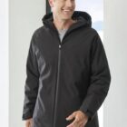 A person wearing a black zip-up hooded jacket stands near a large window. They are smiling and looking to their left. The interior is bright and minimalistic.