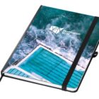 A notebook with a cover featuring an aerial image of a swimming pool by the ocean, with waves crashing. The cover includes the Destination NSW logo. The notebook has a black elastic band and a matching pen holder.
