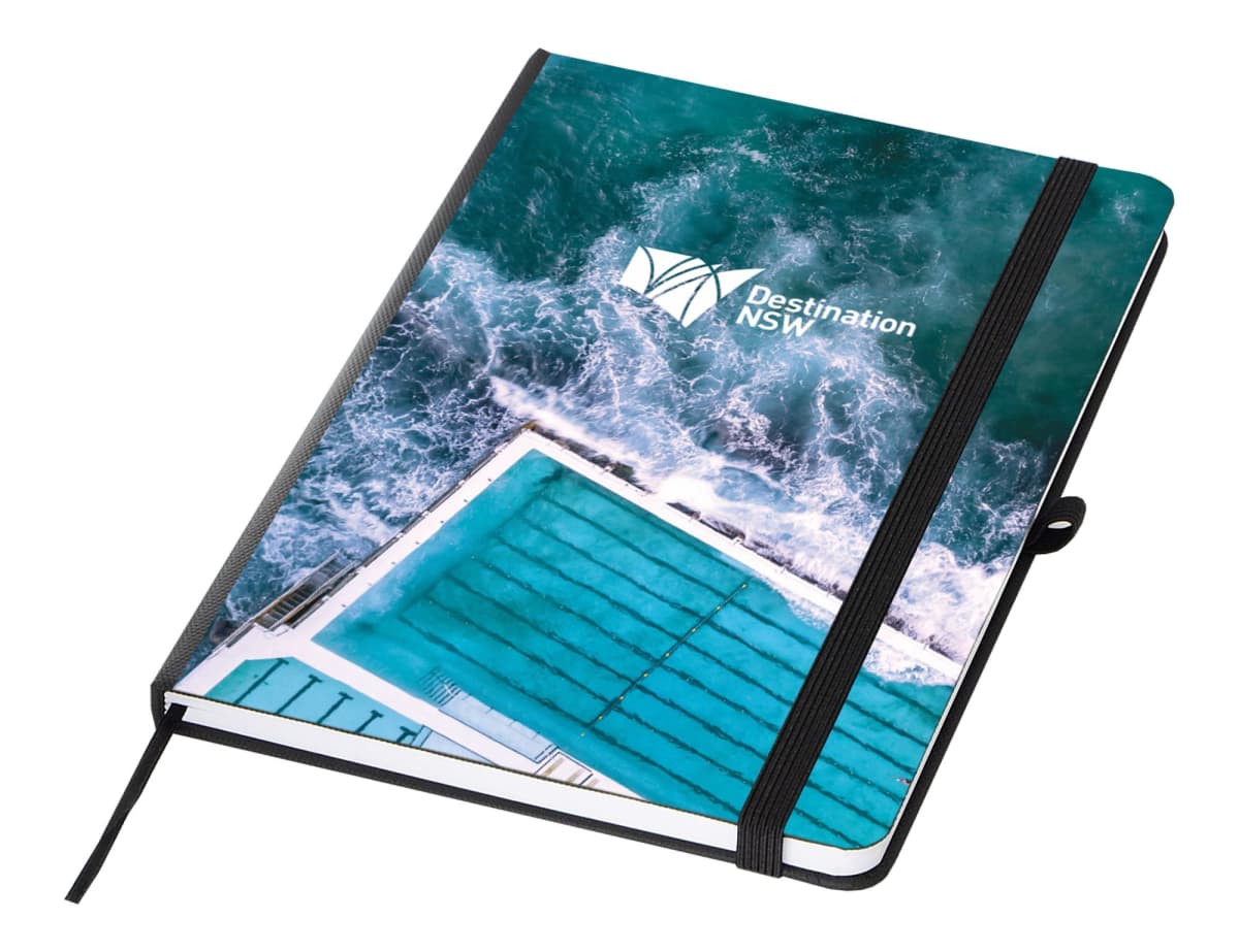 A notebook with a cover featuring an aerial image of a swimming pool by the ocean, with waves crashing. The cover includes the Destination NSW logo. The notebook has a black elastic band and a matching pen holder.