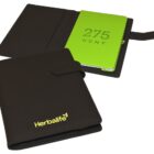 Two black leather folders featuring a green notebook inside. One folder is closed, displaying the Herbalife logo, while the other is open, revealing 275 KENT on the notebook cover. Both folders have a strap closure.