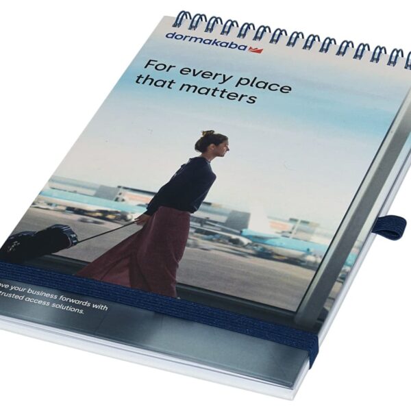 A spiral-bound notebook with a cover image of a person pulling a suitcase at an airport. The text reads dormakaba and For every place that matters. The notebook is secured with an elastic band on the side.