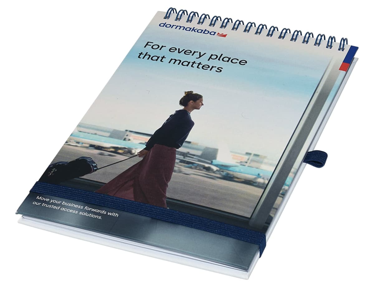A spiral-bound notebook with a cover image of a person walking in an airport, pulling a suitcase. The text on the cover reads, For every place that matters. The notebook features an elastic strap and a pen loop.