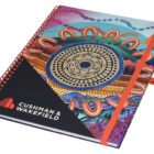 A colorful spiral-bound notebook with an intricate, abstract design featuring circular and wave patterns. The cover displays the Cushman & Wakefield logo in the bottom left corner. The notebook is secured with an orange elastic band.