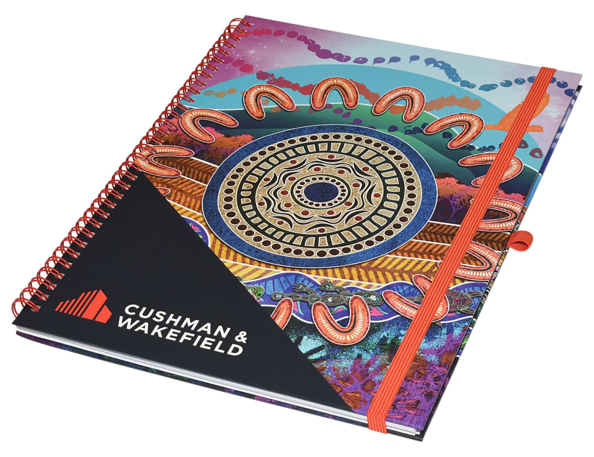 A colorful spiral-bound notebook with an intricate geometric and abstract design on the cover. It features the Cushman & Wakefield logo in the bottom left corner. A red elastic band is attached to the right side for securing the notebook.