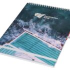 A spiral-bound notebook with a cover featuring an aerial view of a swimming pool next to the ocean. The water is a deep blue-green, and the logo Destination NSW is printed on the top right corner.