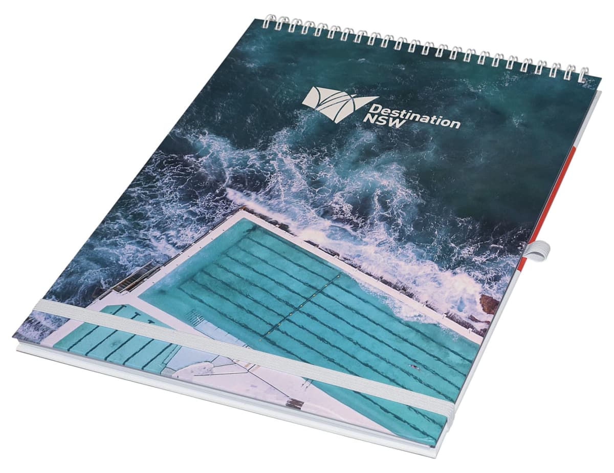 A spiral-bound notebook with a cover featuring an aerial view of a swimming pool next to the ocean. The water is a deep blue-green, and the logo Destination NSW is printed on the top right corner.