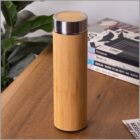 A bamboo insulated water bottle with a stainless steel lid sits on a wooden table. In the background, there are green plants and stacked books, including titles such as Andogynous and Berlin - Born to be Sexy.