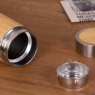A bamboo travel mug lies on its side on a wooden surface, with its stainless steel lid and infuser laid beside it. In the background, a folded newspaper is partially visible.
