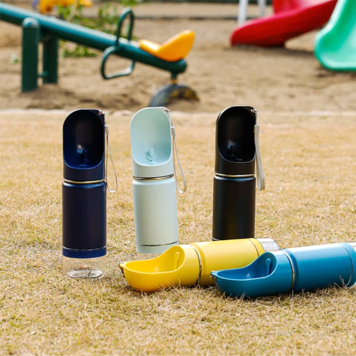 Four portable pet water bottles in black, blue, light blue, and yellow are standing on grass. Each bottle has a detachable drinking cup. Playground equipment is visible in the blurred background.