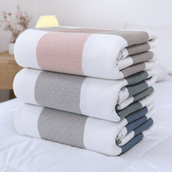A stack of four folded, striped blankets with alternating colors of white, pink, gray, and blue sits on a white bed. A wooden nightstand with a lamp and a small plant is visible in the blurred background.