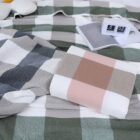 A cozy bed with a checkered blanket in shades of green, beige, and pink. A digital flip clock showing 6:53 sits on the bed along with an open book. A pillow peeks out from the top left corner.