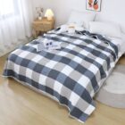 A cozy bedroom with a bed covered in a gray and white checkered blanket. Theres a white flower-shaped cushion and pillows on the bed. A small wooden side table with a lamp and vase, and a window with sheer curtains complete the scene.