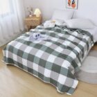 Bedroom with a neatly made bed covered in a green and white checkered blanket. The bed has white pillows and a flower-shaped cushion. A small bedside table with a lamp and decor items is nearby, and natural light filters in through a window with sheer curtains.