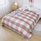 A neatly made bed with a pink and beige checkered blanket in a bright bedroom. A book and plush rabbit are on the bed. A small wooden nightstand with a lamp and decor is beside it, and light streams through sheer curtains.