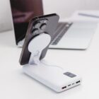 A smartphone is charging wirelessly on a white power bank with a digital display and multiple USB ports. The setup is on a clean white desk with an open laptop in the background.
