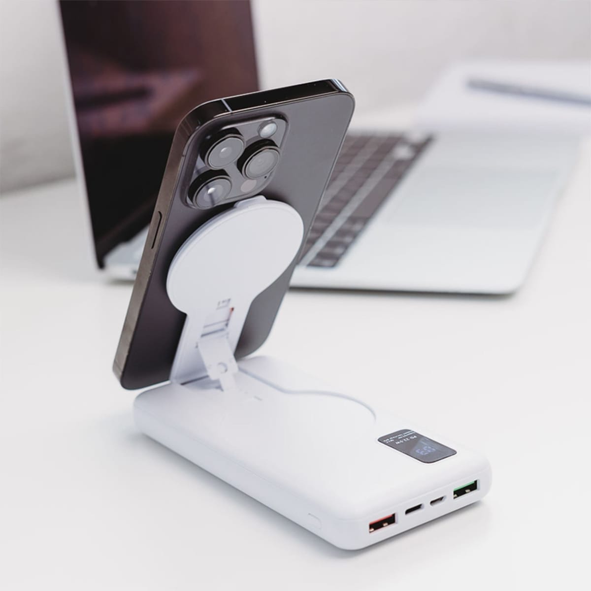 A smartphone is charging wirelessly on a white power bank with a digital display and multiple USB ports. The setup is on a clean white desk with an open laptop in the background.