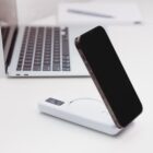A smartphone leaning against a white wireless charging pad on a desk. The charging pad has a small digital display. In the background, part of a laptop and a closed notebook are visible. The scene is bright and minimalist.