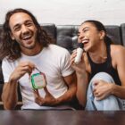 A cheerful man and woman sit on a couch, both laughing. The man holds a colorful Roborock box, while the woman, wearing a black top, holds a microphone. They appear to be enjoying a fun moment together.