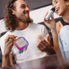A man and a woman are sitting together, smiling and enjoying a karaoke session. The woman is holding a microphone, while the man is holding a compact speaker with colorful lights. They appear to be having fun in a relaxed setting.