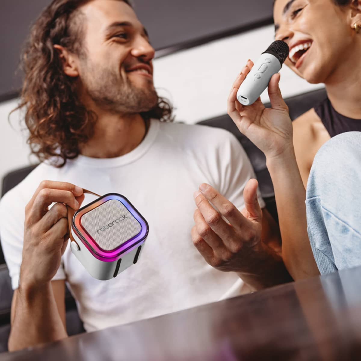 A man and a woman are sitting together, smiling and enjoying a karaoke session. The woman is holding a microphone, while the man is holding a compact speaker with colorful lights. They appear to be having fun in a relaxed setting.